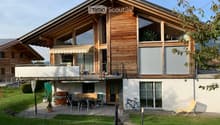 Two-story wooden house with large windows, balcony, outdoor seating area, and garden.