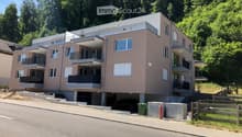 Modern multi-story residential building with balconies, large windows, and a parking area at the base.