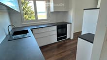 Modern kitchen with white cabinets, dark countertops, wooden flooring, and a large window overlooking greenery.