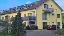 Two-story yellow house with red-tiled roof, multiple windows, balconies, and a driveway with a parked car.