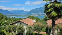 A serene garden view with a small white house, surrounded by lush greenery and palm trees, overlooking a lake and distant mountains.