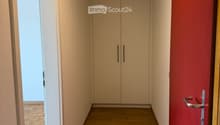 A narrow hallway with wooden flooring, light-colored walls, and a built-in closet with double doors. The hallway features a red accent wall on one side and a partially open door leading to another room.
