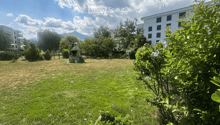 A spacious, well-maintained garden area with green grass, trees, shrubs, and a small stone fireplace in the background. The area is bordered by a modern building and features a clear, partly cloudy sky.