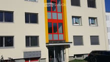 Two-story building with light beige walls, red and yellow entrance door, multiple windows with blinds, and a small parking area with bicycles and a car.