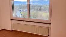 Empty room with wooden flooring, large windows offering a scenic mountain and lake view, radiator, and wooden ceiling beams.