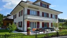 Two-story house with white exterior, red shutters, and a balcony on the upper floor. The front yard features a well-maintained lawn, a black wrought-iron fence, and outdoor seating including a sofa, coffee table, and dining table with chairs.