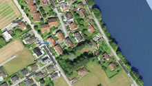 Aerial view of a residential area with multiple houses, gardens, and surrounding fields. The houses are clustered along streets, with some near a body of water.