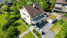 Aerial view of a two-story house with a brown tiled roof, white walls, and a spacious balcony. The property is surrounded by a well-maintained lawn and trees, with neighboring houses visible in the background.