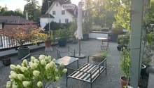 A well-maintained garden area with gravel ground, potted plants, a metal bench, a small table, and chairs, with an umbrella and surrounding greenery.