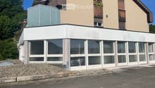 Modern two-story building with a flat roof and large windows, featuring a raised ground floor and a balcony with plants.