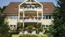 Two-story house with a brown tiled roof, white exterior, multiple windows, and balconies adorned with flower boxes and greenery.
