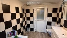 Modern bathroom with checkered black and white tiles, wooden flooring, bathtub, sink, toilet, shower, and a small stool. Includes a black bath mat with white text and a small plant on the sink.