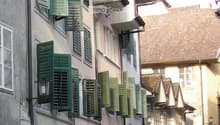 A narrow cobblestone street lined with multi-story buildings featuring green shutters and classic architectural details.