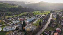 Aerial view of a suburban neighborhood with residential buildings, roads, and surrounding green fields and hills.