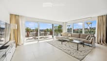 Modern living room with large windows and a balcony view, featuring a beige sofa, glass coffee table, floor lamp, TV stand, and dining area with chairs and table.