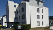 Modern, multi-story white building with rectangular windows and a flat roof, surrounded by a small landscaped area with hedges and a trash bin.