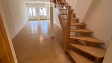 Spacious hallway with wooden flooring, a staircase with wooden steps and a metal railing, and a view into an adjacent room with large windows and a radiator.