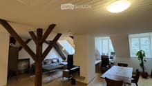Open-plan living and dining area with wooden floors, exposed wooden beams, large windows, sofa, coffee table, dining table, chairs, and a plant.