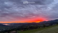A panoramic view of a rural landscape at sunset, featuring rolling green hills, scattered houses, a body of water, and distant mountains under a dramatic sky.