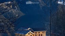 A modern wooden house with a dark roof, situated in a snowy mountainous landscape with bare trees in the foreground.