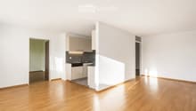 Open-plan unfurnished room with light wooden flooring, white walls, and a small kitchen area featuring cabinets, sink, and dishwasher.
