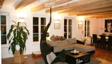 Living room with exposed wooden beams, large windows, a grey sectional sofa, a black wicker hanging chair, a coffee table, and a dining area with chairs and a table.