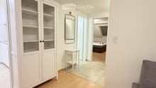 A modern, minimalist living room with light wooden flooring, white walls, a grey sofa, a white cabinet with glass doors, a small white side table, and a mirror on the wall. The room is connected to another space with a bed visible in the background.