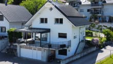 A modern two-story house with white exterior walls, grey tiled roof, large windows, and a covered patio area with seating. The house features a garage and is surrounded by greenery and neighboring houses.