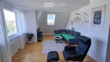 Modern living room with wooden flooring, large windows, a green sofa, a black leather chair, a coffee table, a small plant, and artwork on the walls.