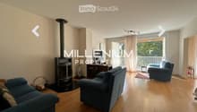 Spacious living room with wooden flooring, fireplace, large windows, and balcony access. Features a blue sofa, armchair, and small table.