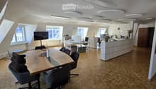 Bright, modern office space with wooden flooring, large windows, a long wooden conference table with chairs, a TV screen, desks, cabinets, and decorative plants.