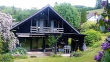 Two-story house with a dark wooden exterior, large windows, and a balcony on the upper level. The surrounding area includes a well-maintained garden with flowering trees and shrubs.