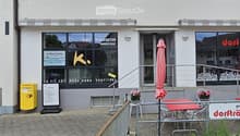 Facade of a commercial building with large windows, signage for "Konfi Manufaktur Markus Kunz" and "LA VIE EST DURE SANS CONFITURE", outdoor seating area with tables and chairs, and a yellow post box.