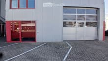 Modern building with a large garage door and a glass entrance. The facade is grey with red trim around the windows and doors. The ground is paved with bricks and marked parking spaces.