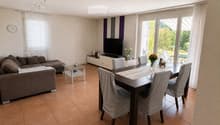 Modern living and dining area with a brown sectional sofa, glass coffee table, dark wooden dining table with four chairs, flat-screen TV on a white stand, and decorative vases on the table and TV stand.