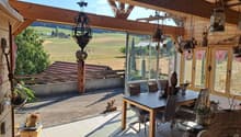 Bright, wooden-framed balcony with large windows offering a scenic countryside view. Features a dining table with chairs, hanging decorations, and potted plants.