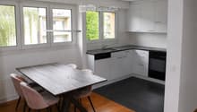 Modern kitchen with white cabinets, black countertops, a wooden dining table with four pink chairs, and large windows offering a view of greenery.
