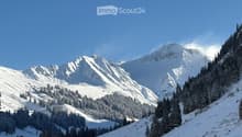 Snow-covered mountain landscape with dense evergreen forest, clear blue sky, and ski lift visible in the distance.