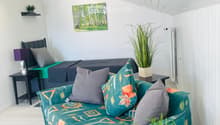 A modern, well-lit room with a green sofa, a bed with dark bedding, a nightstand, a small table with a potted plant, and a picture frame on the wall.