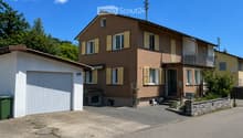 Two-story house with brown brick facade, white garage, and balcony on the upper floor. The house features multiple windows with shutters and a small garden area in front.