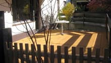 Wooden deck with a small round table, surrounded by a wooden fence, with a view of the house and garden.
