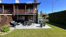 Well-maintained garden with a patio area featuring a sofa, table, chairs, and a grill. The house has a wooden balcony and a well-kept lawn.