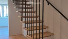 Wooden staircase with black metal balustrade, light wooden steps, and a handrail, set against light-colored walls and wooden flooring.