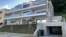 Modern two-story house with large glass balconies, raised ground floor, and garage. The house features a beige facade, multiple large windows, and a landscaped hillside entrance.