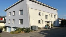 Modern two-story house with light grey siding, multiple windows, and a small garage. There are bicycles and a scooter parked near the entrance, along with some greenery and a trash bin.