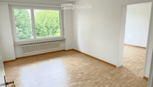Empty room with wooden flooring, white walls, large window with view of greenery, radiator below window, and a door visible on the left side.