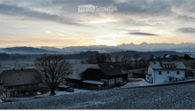 Aerial view of a house with a tiled roof, surrounded by a snow-covered yard and trees, with a scenic mountain backdrop at sunset.