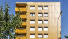 A multi-story residential building with yellow balconies and white-framed windows, surrounded by greenery and a clear blue sky.