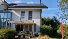 Two-story house with solar panels on the roof, large windows, and a covered patio area with outdoor furniture.