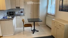 Compact kitchen with light wood cabinets, granite countertop, microwave, sink, small dining table with chair, and vase on counter.